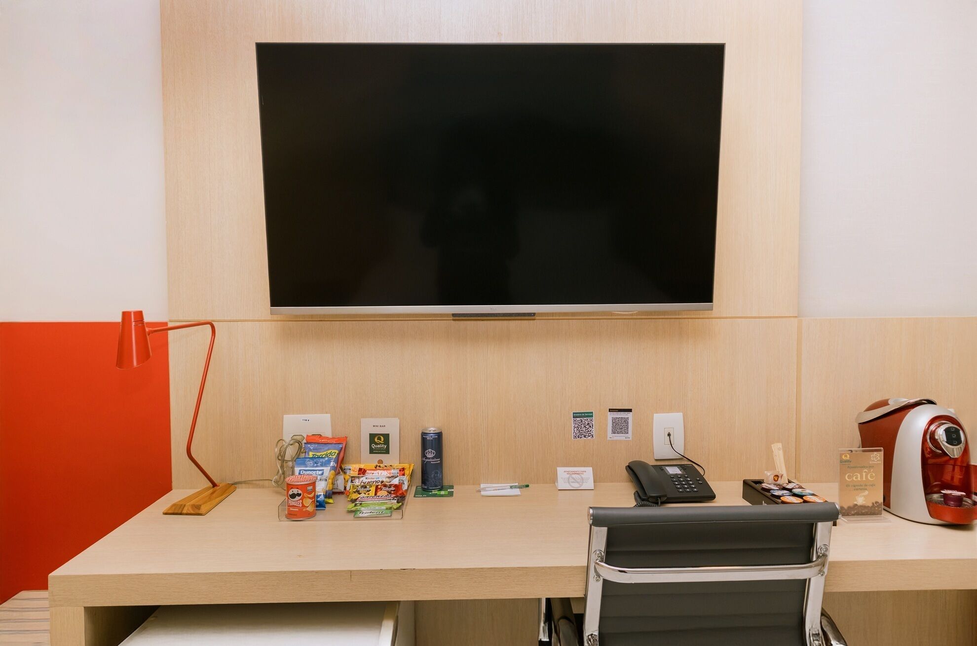 A hotel room with a large flat-screen TV mounted on the wall, a desk with various amenities including snacks, a phone, and a kettle.