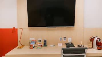 A hotel room with a large flat-screen TV mounted on the wall, a desk with various amenities including snacks, a phone, and a kettle.