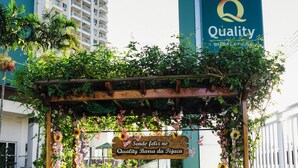 Una pérgola de madera con un banco, rodeada de abundante follaje y flores, con un letrero que dice "Sendo feliz no Quality Barra da Tijuca".