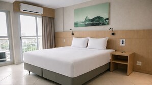 Habitación de hotel con una cama grande, una mesita de noche, aire acondicionado en la pared, una ventana con cortinas y un cuadro de un paisaje urbano.
