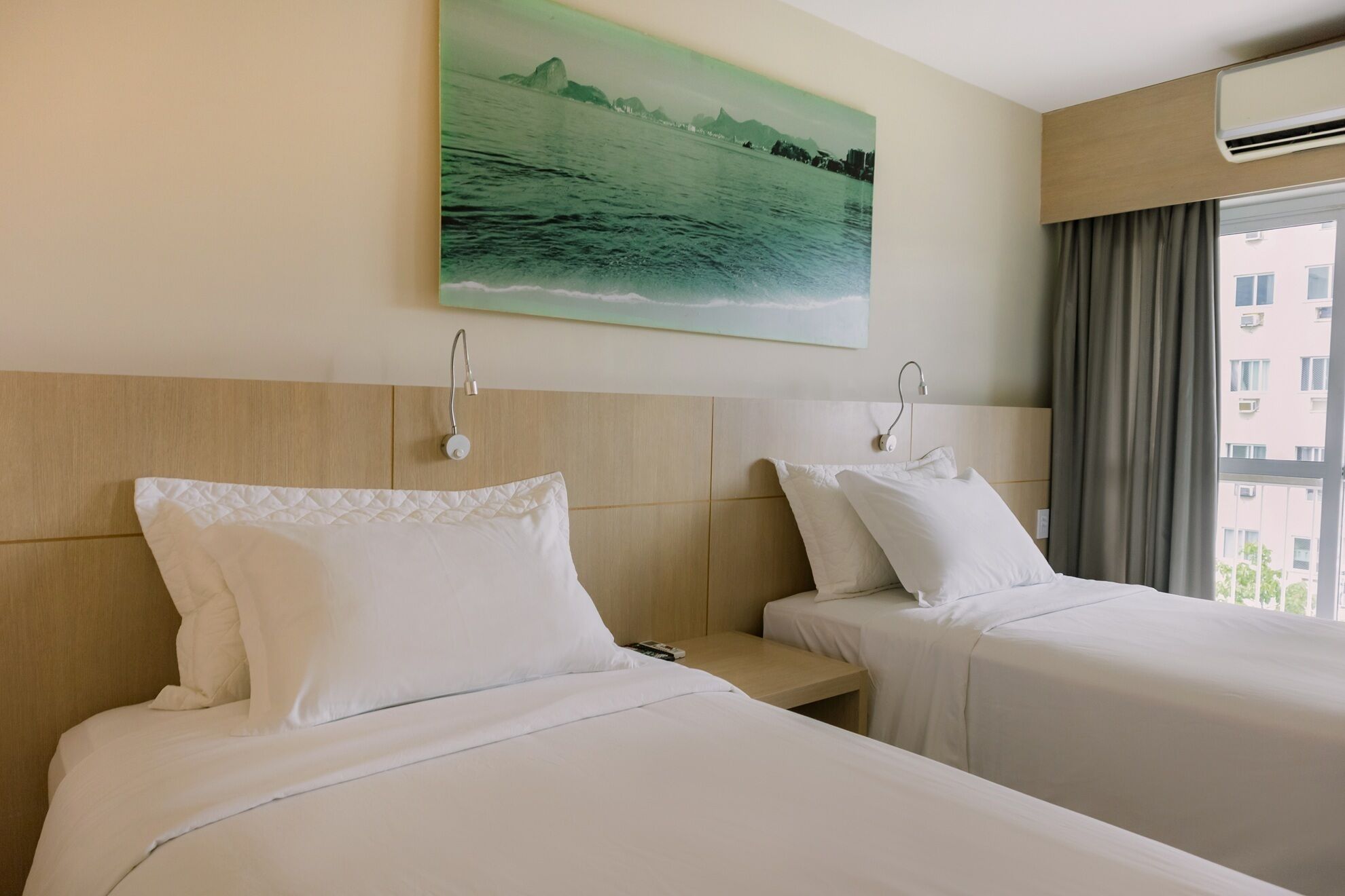 Habitación de hotel con dos camas, un cuadro de una escena costera, una ventana con cortinas y un equipo de aire acondicionado.