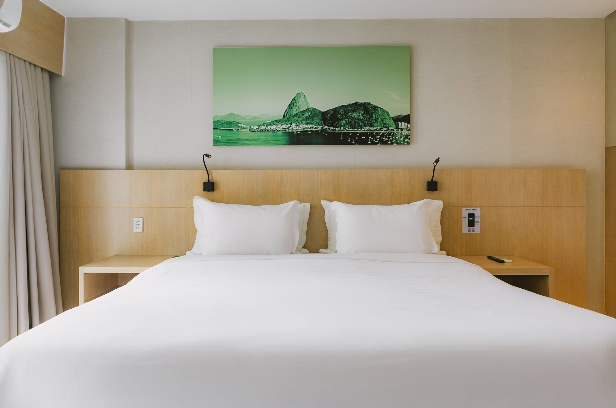 A hotel room with a large bed, two bedside tables, a nightstand, a wall-mounted lamp, and a painting of a landscape.