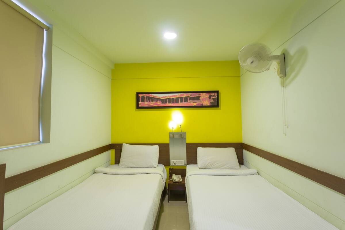 A hotel room with two beds, a bedside table with a phone, a wall-mounted fan, and a painting above the bed.