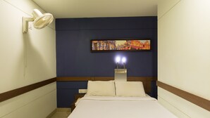 A hotel room with a bed, a wall-mounted fan, a painting, and a bedside table with a lamp. - Ginger Chennai Vadapalani (Chennai)