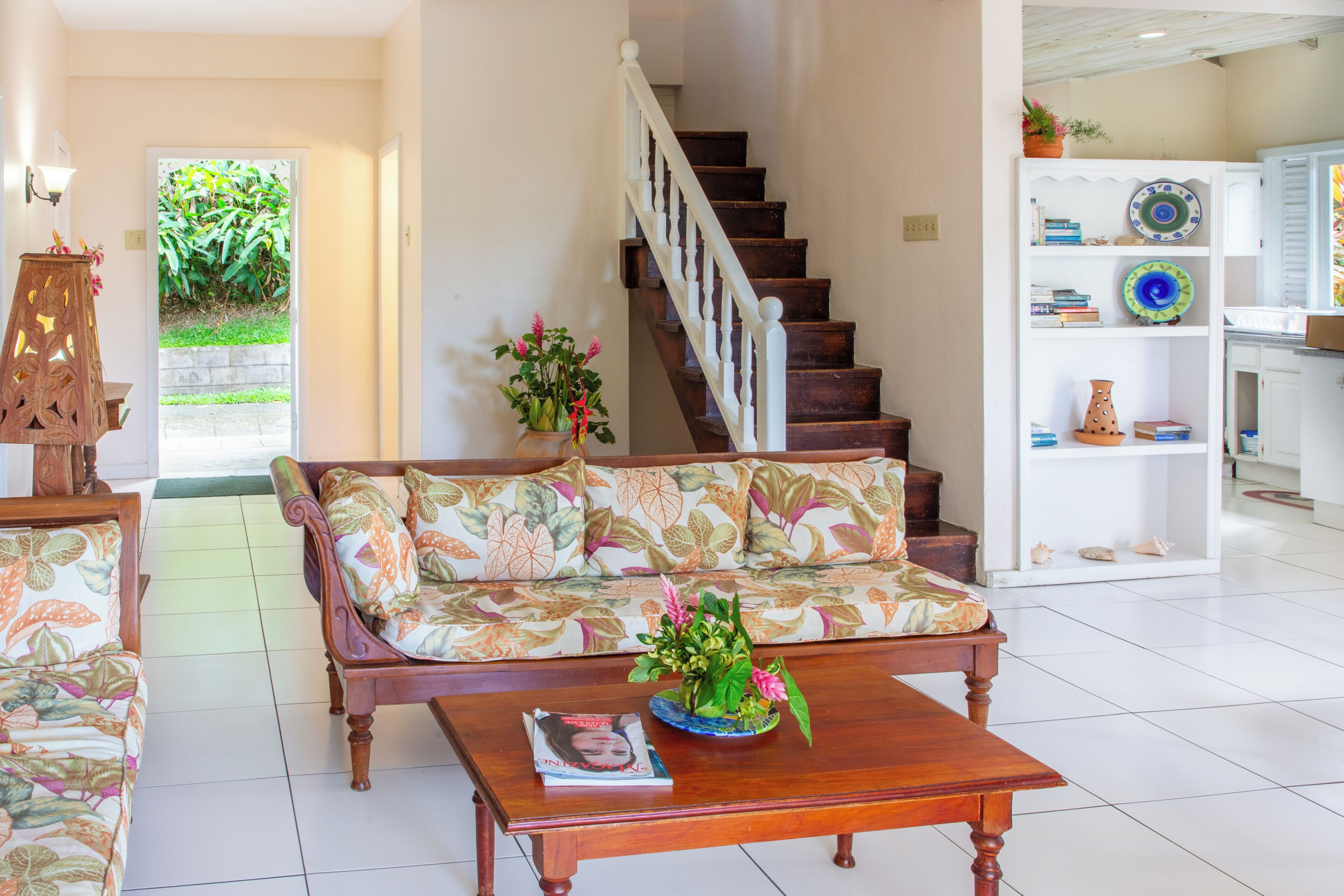 A living room with a wooden staircase, a sofa with floral cushions, a coffee table with a magazine, and a bookshelf with decorative plates.