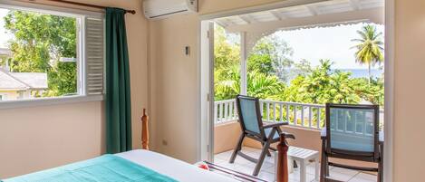 Une chambre avec un lit, une chaise et un balcon donnant sur un espace verdoyant et une étendue d’eau.