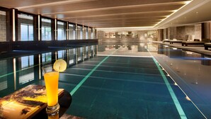 A modern indoor swimming pool with a glass barrier, lounge seating, and a glass of orange juice on a table. - Banyan Tree Tianjin Riverside (Tianjin)