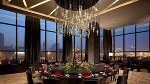 A dining area with a round table set for six, surrounded by six chairs. A large chandelier hangs from the ceiling, and the room features large windows with a cityscape view. - Banyan Tree Tianjin Riverside (Tianjin)