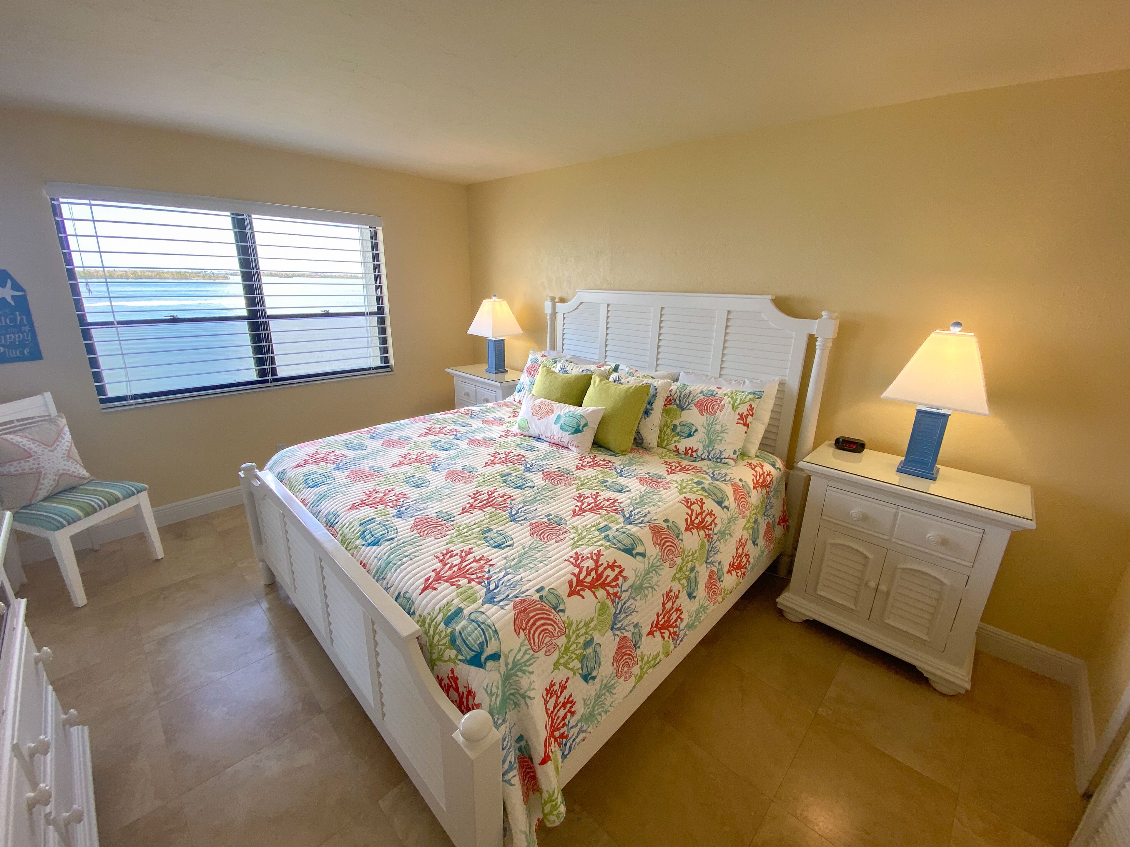 A bedroom with a bed, two bedside tables with lamps, a chair, a window with blinds, and a view of the sea.