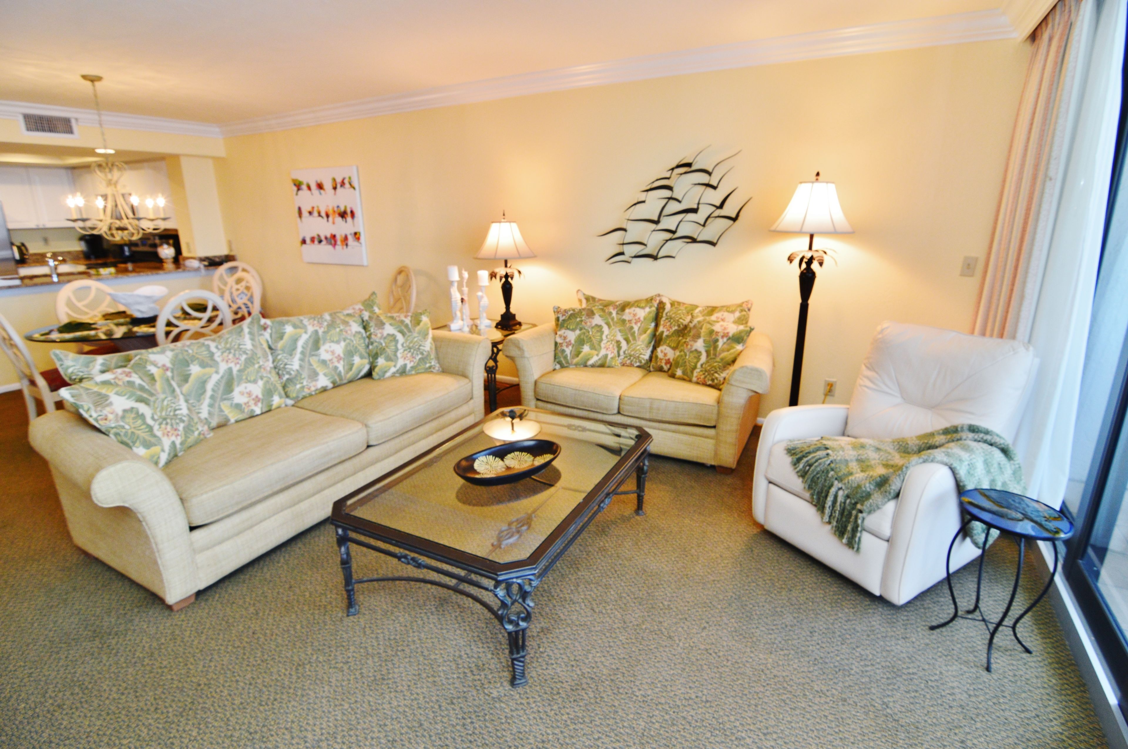 A living room with a beige sofa, a white armchair, and a coffee table. There is a chandelier, wall art, and a lamp on a side table.