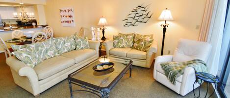 A living room with a beige sofa, a white armchair, and a coffee table. There is a chandelier, wall art, and a lamp on a side table.