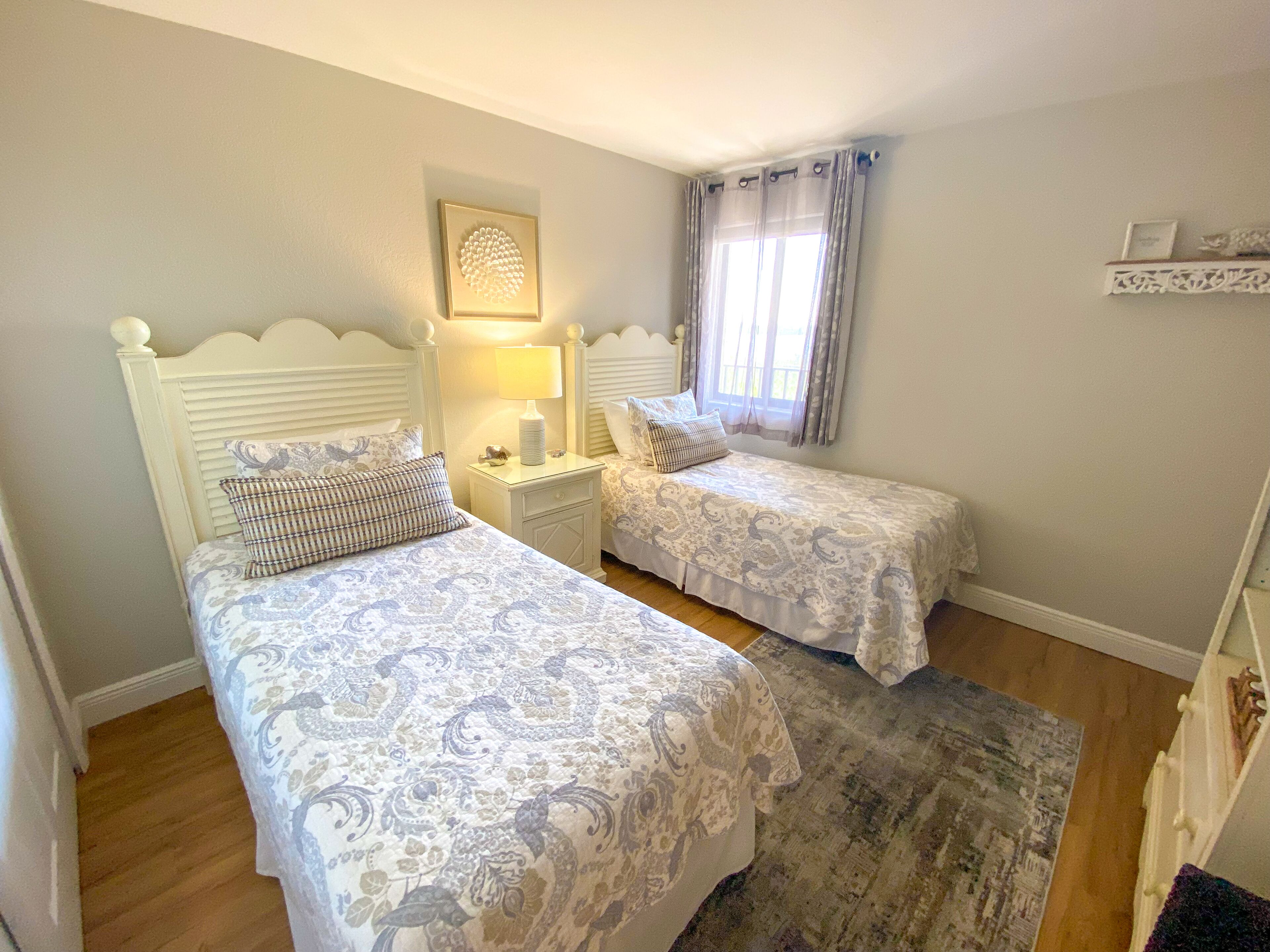 A bedroom with two beds, a nightstand, a wall-mounted lamp, and a window with curtains.