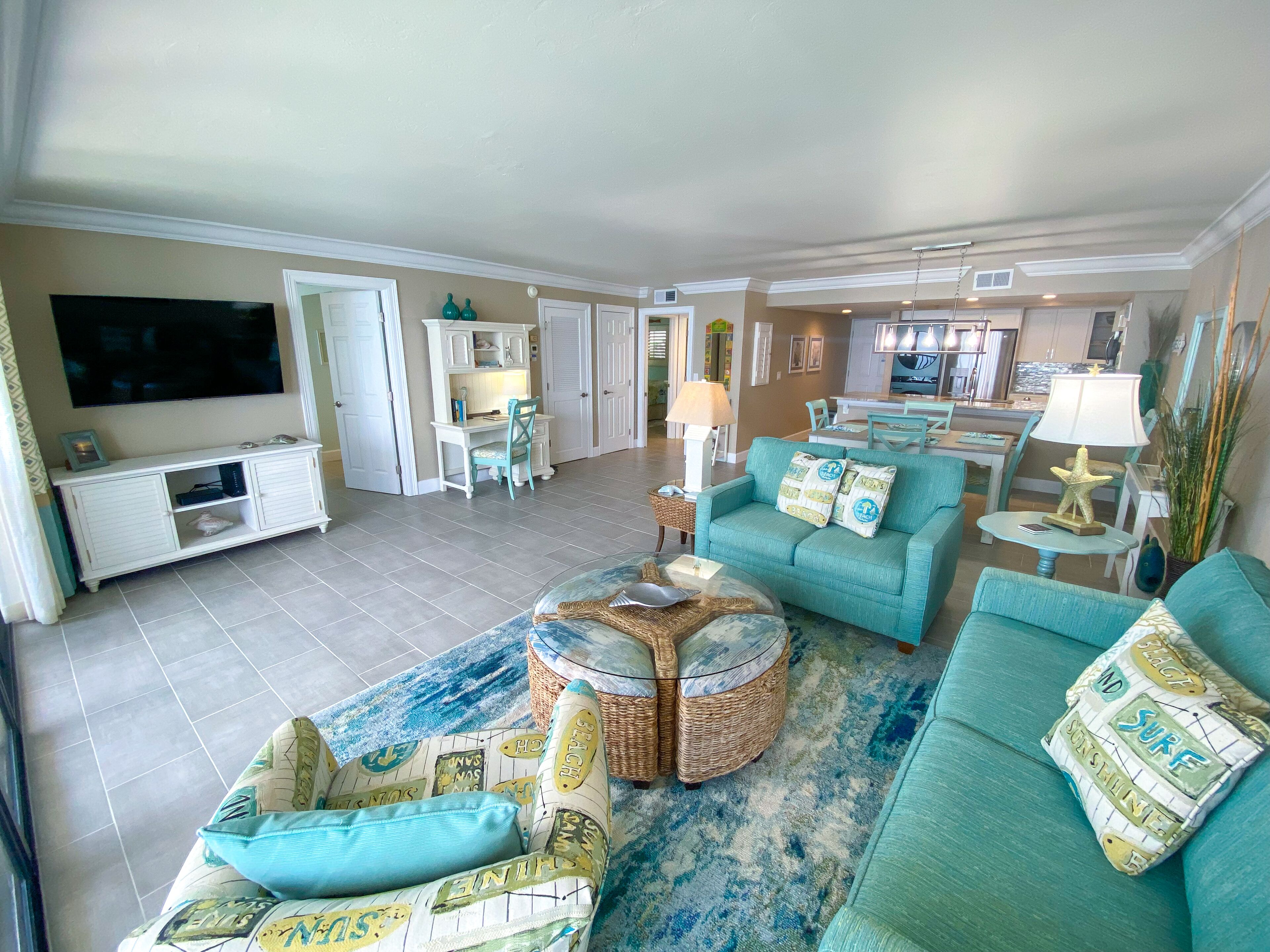 A spacious living room with a large TV, teal sofas, and a dining area with a table and chairs.