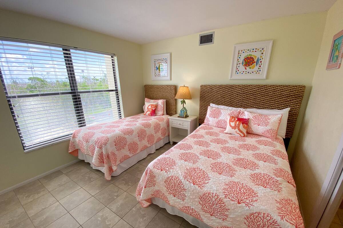 A bedroom with two beds, a window with blinds, a nightstand with a lamp, and two framed pictures on the wall.