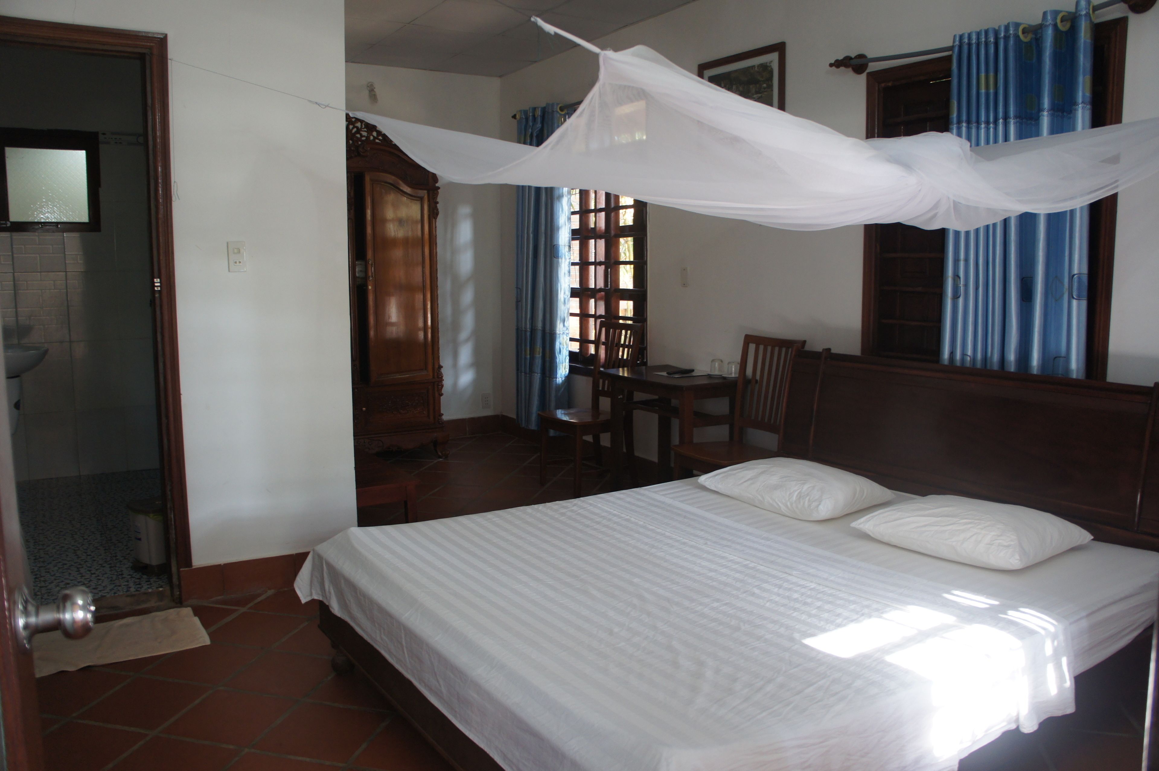 A bedroom with a bed, a wooden headboard, a desk with chairs, and a window with curtains.