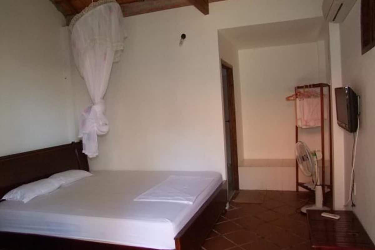 A bedroom with a bed, a wooden headboard, a white curtain, a small table, and a wall-mounted TV.