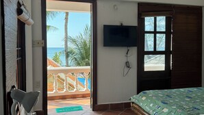 Ett rum med en säng, en tv som är monterad på väggen, ett bord med en stol och utsikt över havet genom en öppen dörr.