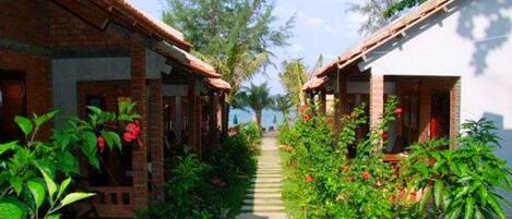 A pathway leading to a row of traditional-style buildings with brick walls and tiled roofs, surrounded by lush greenery and flowering plants.