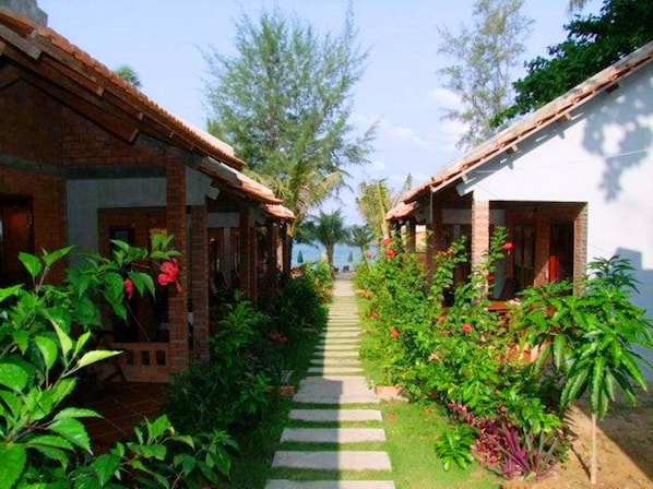 A pathway leading to a row of traditional-style buildings with brick walls and tiled roofs, surrounded by lush greenery and flowering plants.