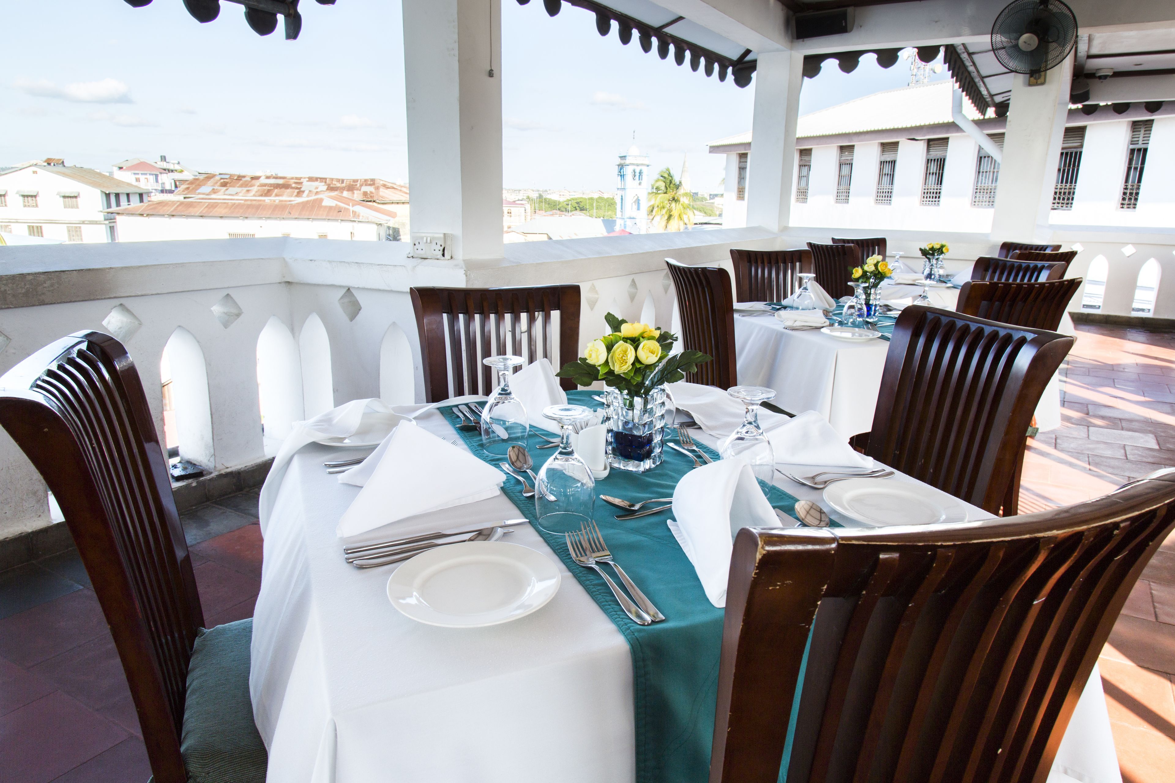 A dining area with tables set for four, each with white tablecloths, chairs, and a vase of yellow flowers.