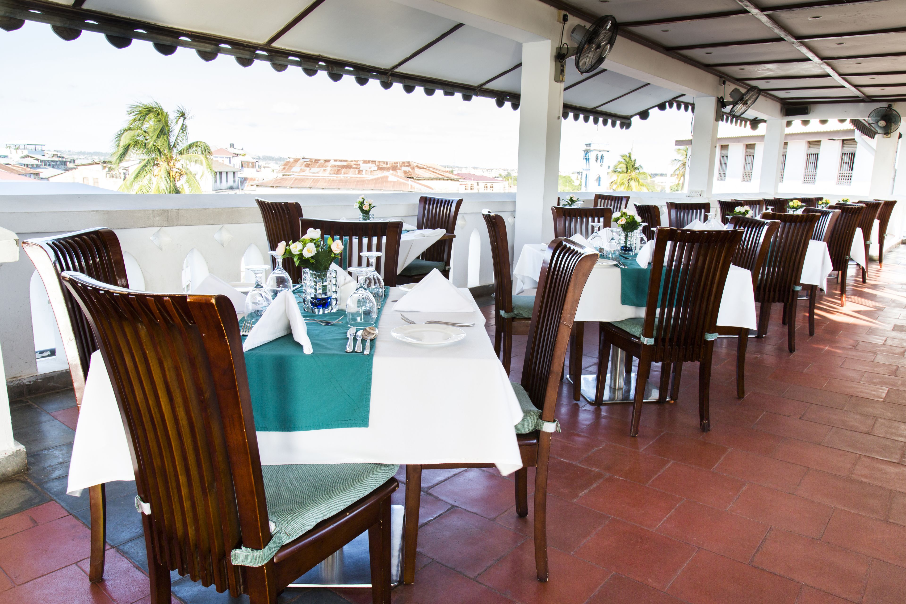 A restaurant with outdoor seating, tables set with white tablecloths and wooden chairs, and a view of the cityscape.