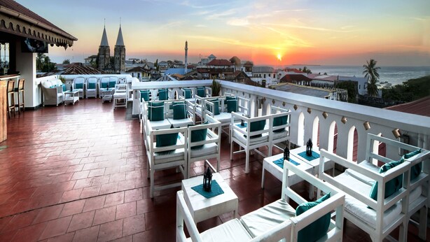 A rooftop terrace with white furniture and a view of a historic church and sunset over the sea. - Maru Maru Hotel (Zanzibar Town)