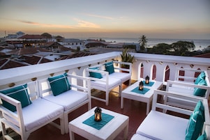 Terraza en la azotea con mobiliario blanco, cojines color turquesa y faroles, con vistas a una ciudad costera.