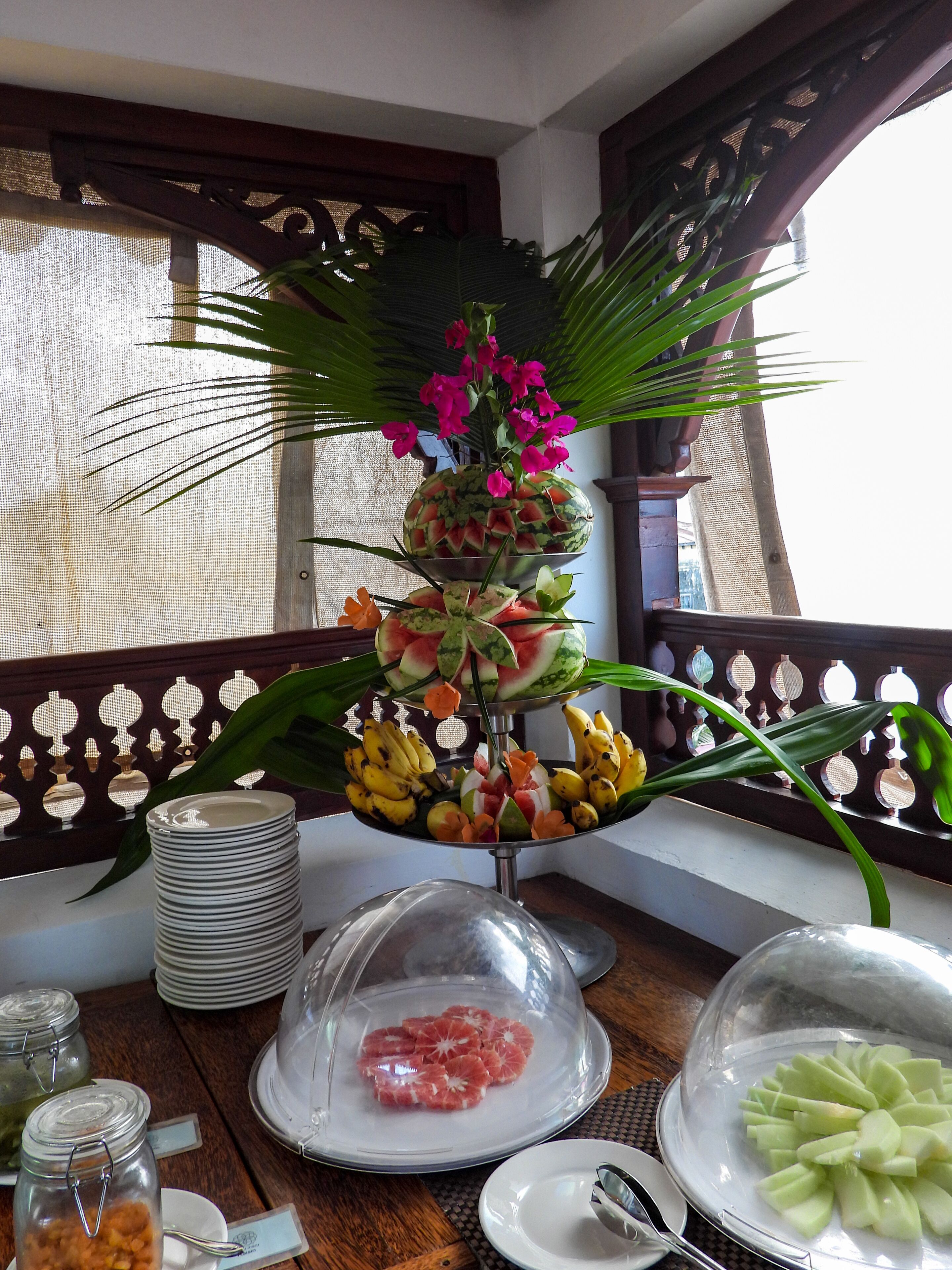 Une table agrémentée d’une pièce maîtresse décorative composée d’une pastèque, de fleurs et de fruits, entourée d’assiettes et recouverte d’un cloche de verre protégeant les aliments.