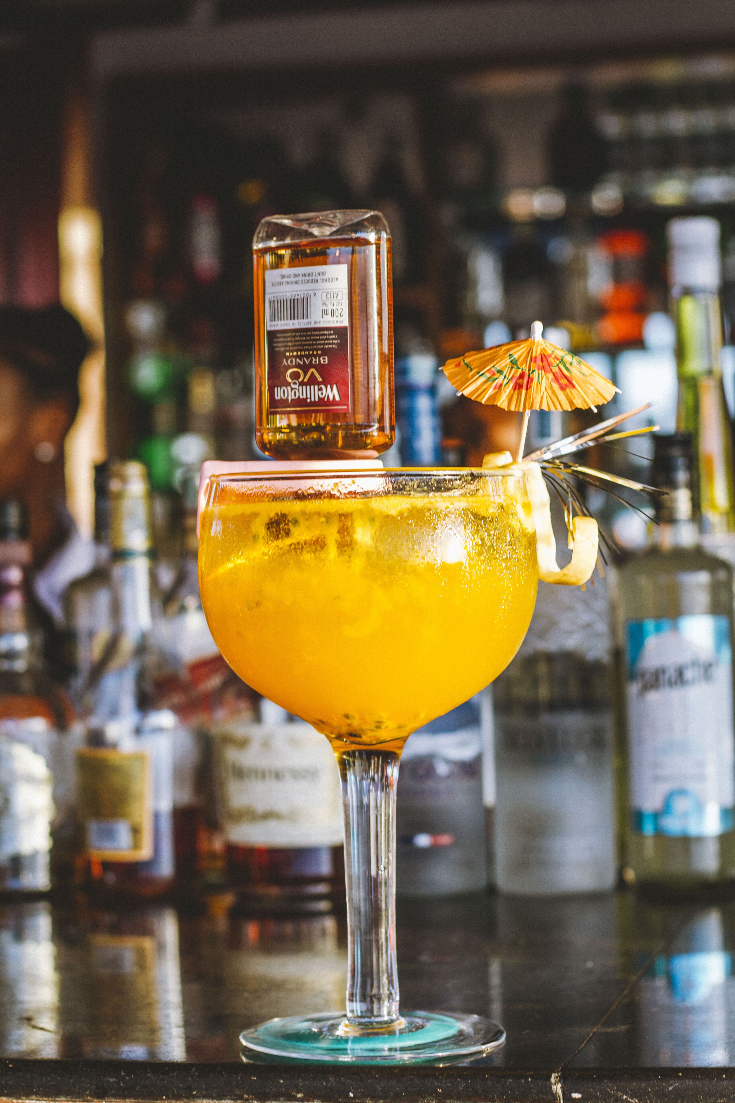 Um cocktail com uma garrafa de vodka e um guarda-chuva na borda, apoiado em um balcão de bar com diversas garrafas ao fundo.