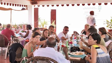 A group of people dining at an outdoor restaurant with wicker furniture and potted plants.