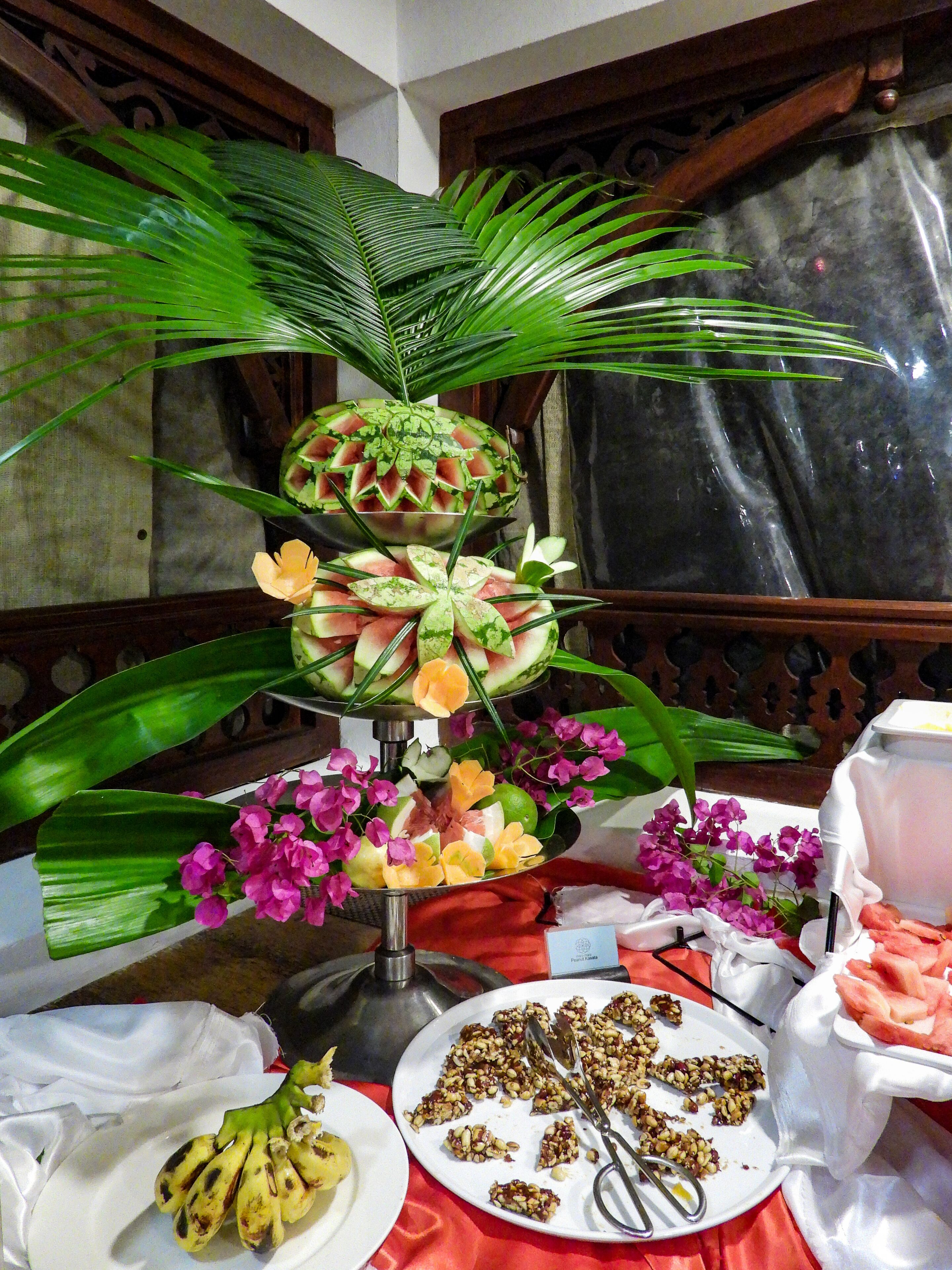 A table with a fruit arrangement, including a watermelon centerpiece, surrounded by plates of food and utensils.