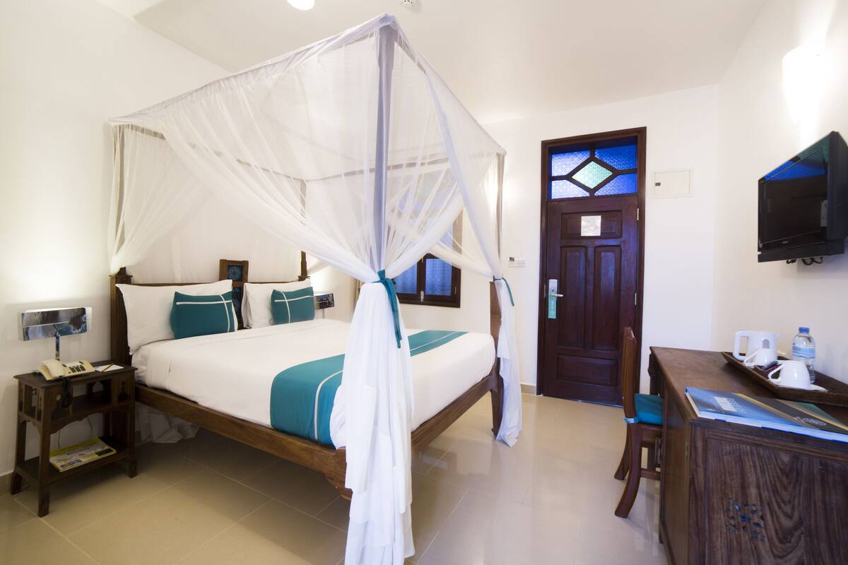 Une chambre d’hôtel avec un lit à baldaquin, un bureau en bois, une chaise, un téléviseur fixé au mur et une porte ornée de vitraux.