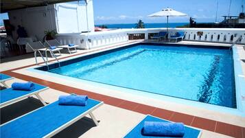A rectangular swimming pool with a clear blue water surface, surrounded by sun loungers with blue towels, and a view of the ocean in the background.