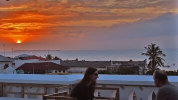A sunset view from a rooftop terrace with a palm tree and a body of water in the background.