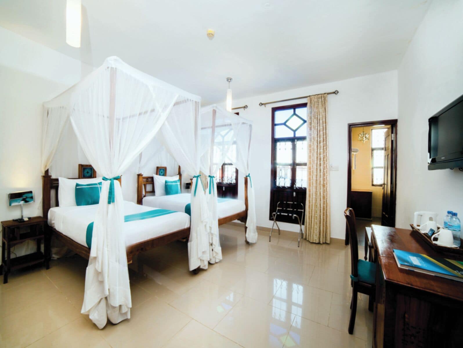A hotel room with two beds, a desk, a chair, a TV, and a large window with curtains.