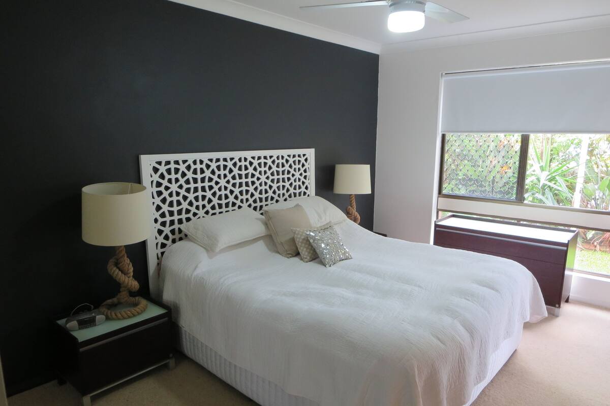 A neatly made bed with white linens, two bedside tables with lamps, and a window with a view of greenery.