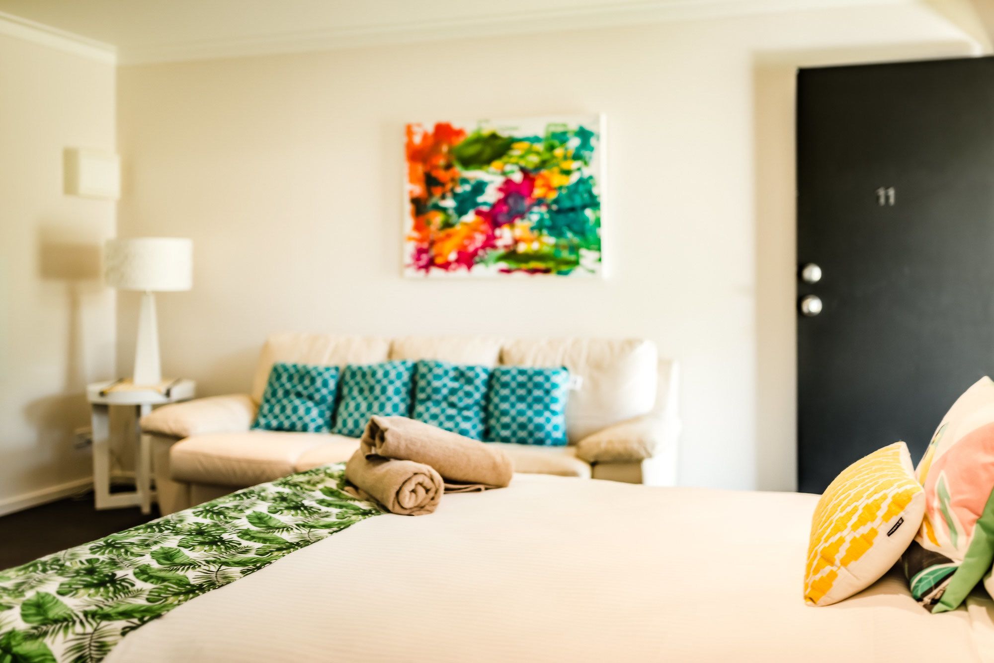 Une chambre d’hôtel avec un lit, une table de chevet, une lampe, un tableau abstrait aux couleurs vives et une porte portant un numéro.