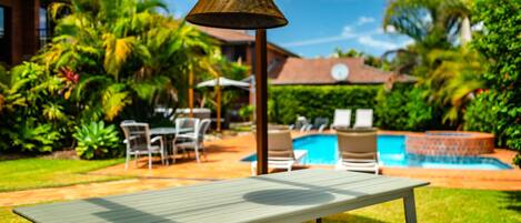 Zona de piscina con mesa y sillas, rodeada de abundante vegetación y un cielo azul despejado.