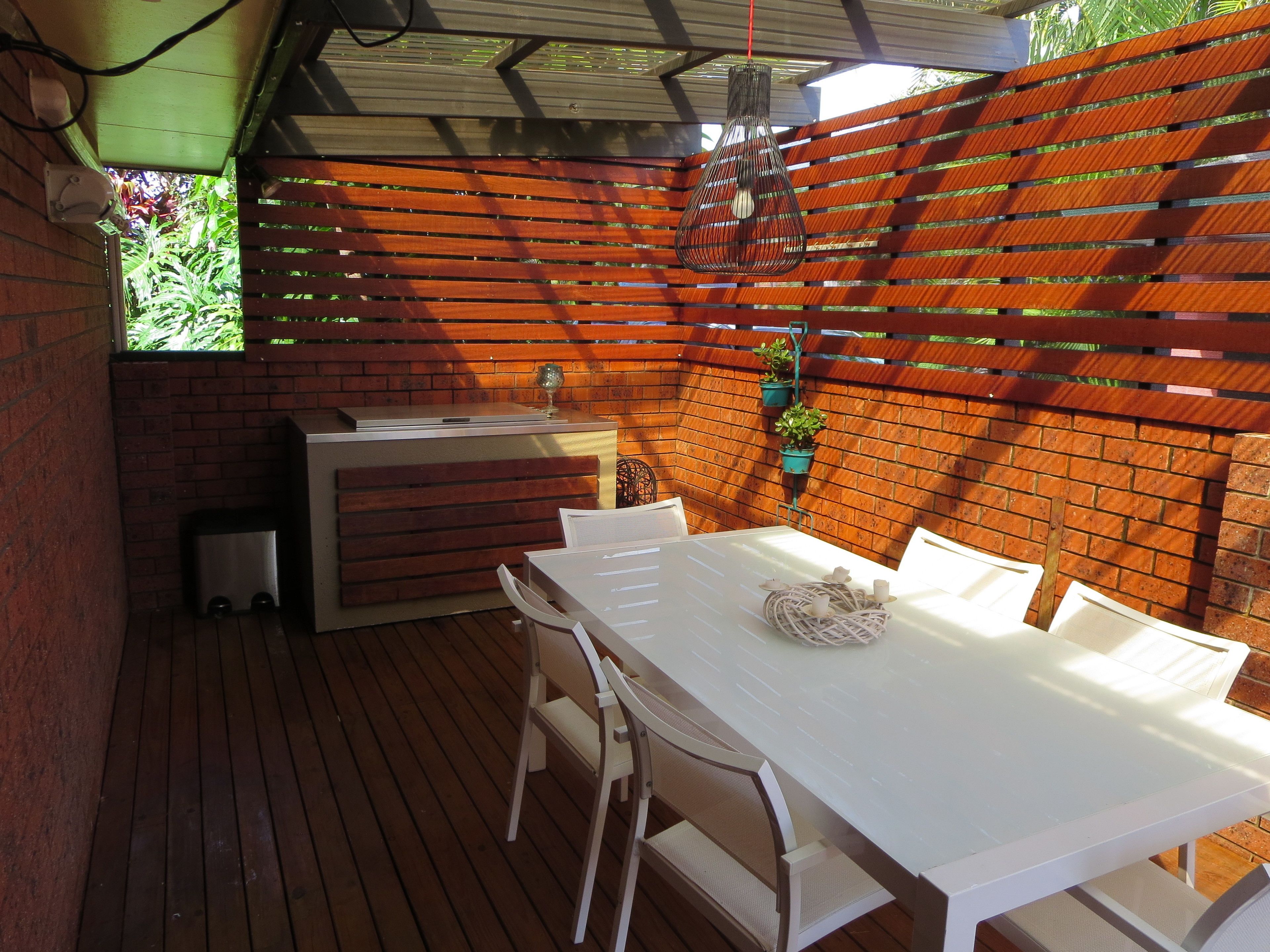 Een terras buiten met een houten dek, een witte tafel en stoelen. Achtergrond is een bakstenen muur en een houten omheining.