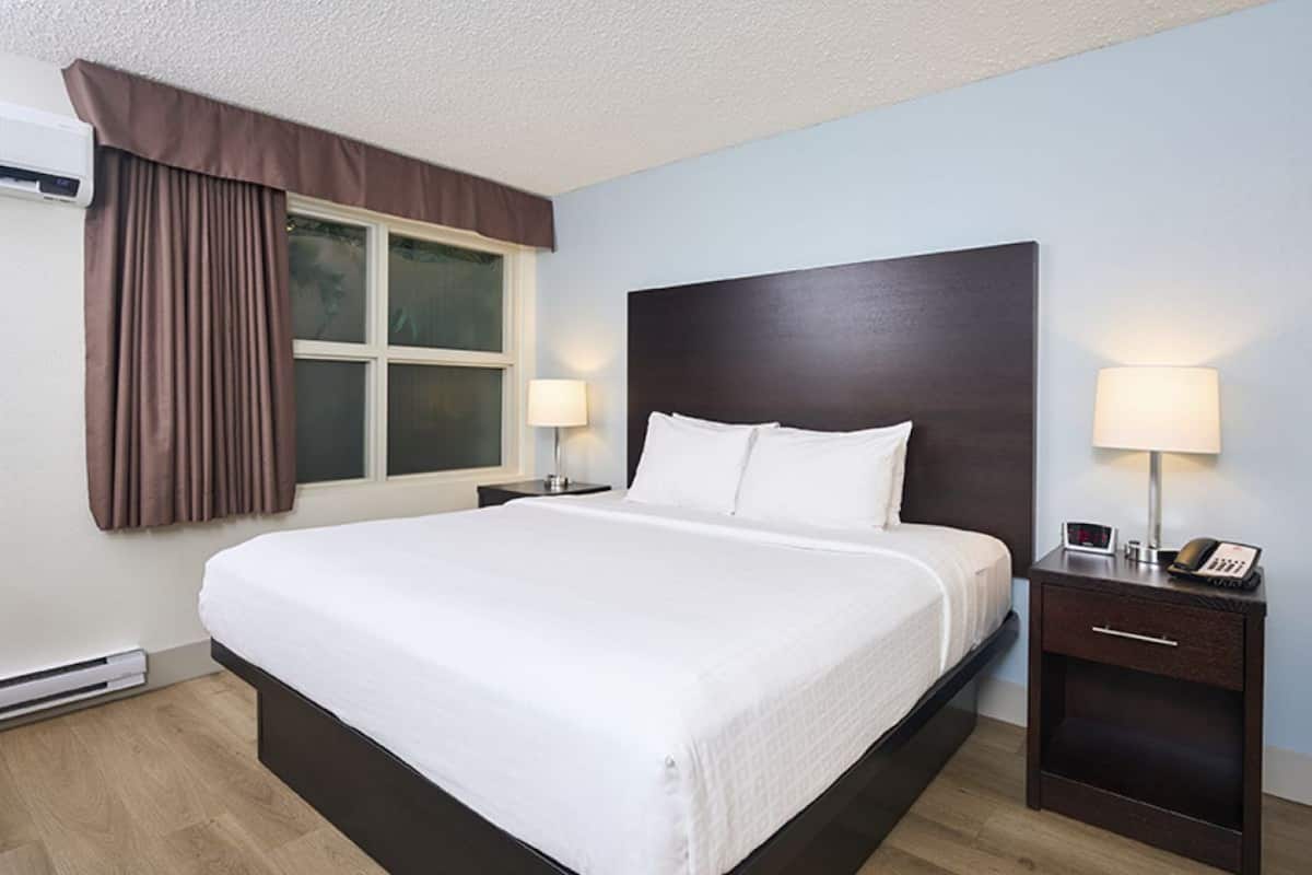 A hotel room with a large bed, two bedside tables with lamps, a window with curtains, and a thermostat on the wall.
