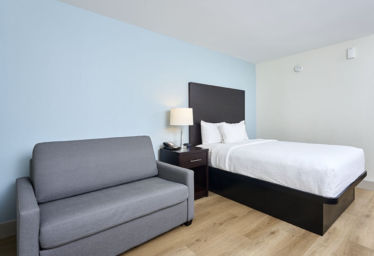 Une chambre d’hôtel avec un lit, un canapé gris, une table de chevet avec une lampe et un plancher en bois.