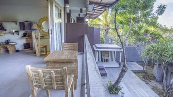 A covered outdoor area with a wooden table and chairs, a small kitchen area, and a view of a garden with trees and plants.