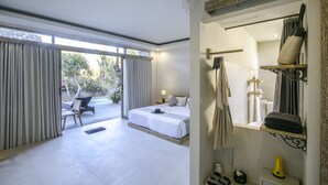 A modern hotel room with a large bed, a sitting area with a chair, a small table, and a bathroom with a mirror and towel rack. - KajaNe Tulamben (Kubu)