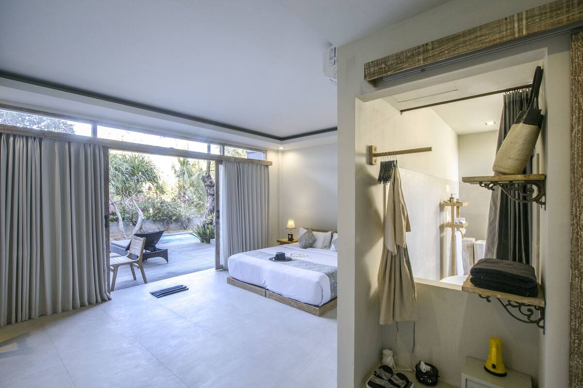 A hotel room with a large bed, a chair, a small table, a mirror, and a view of the outdoors.