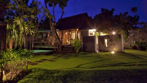 A well-lit house with a thatched roof, surrounded by lush greenery and a neatly maintained lawn. - KajaNe Tulamben (Kubu)