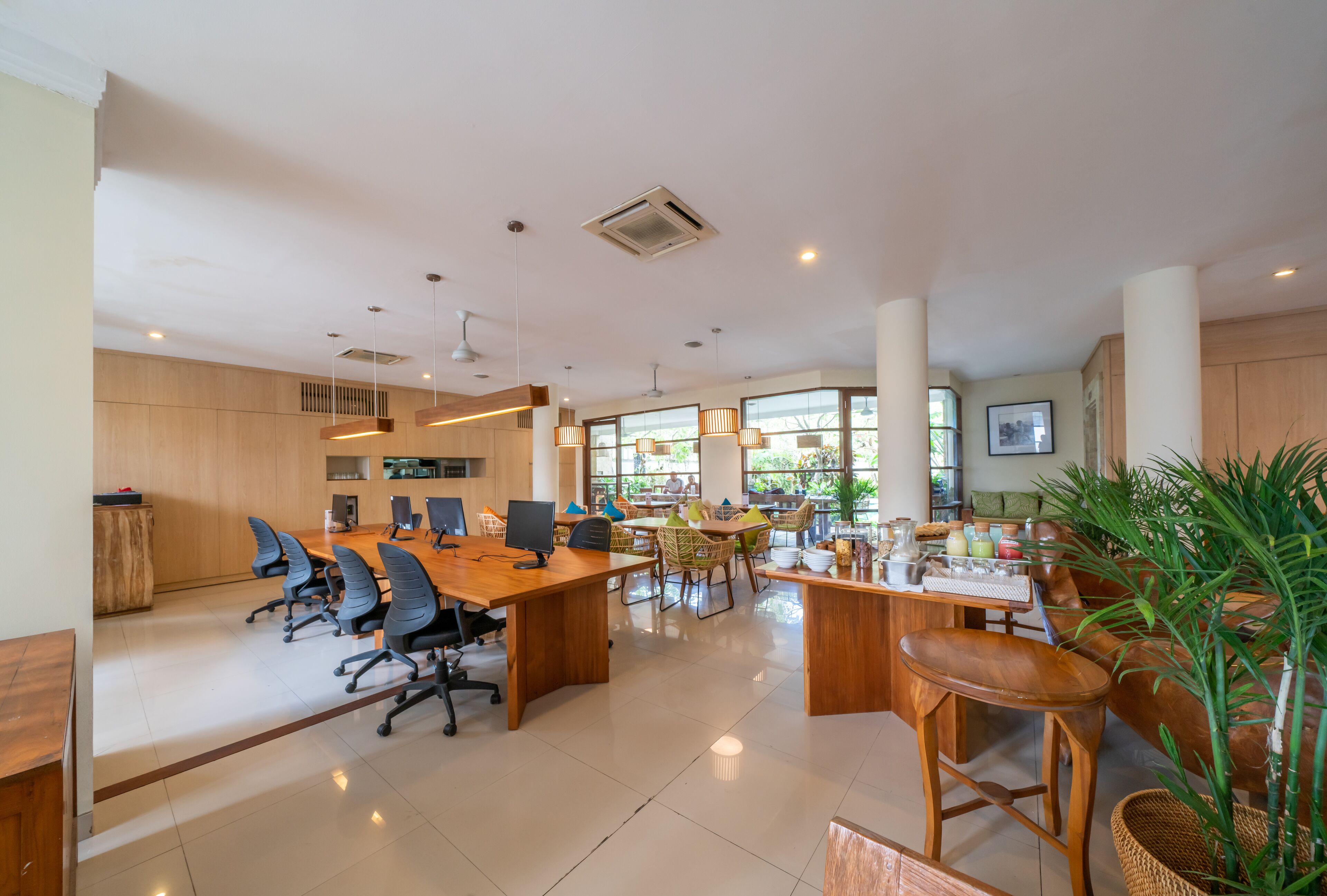 A spacious office with multiple desks, chairs, and a large meeting table. There are several plants and a television mounted on the wall.