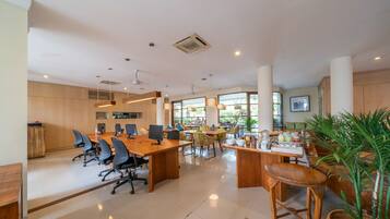 A spacious office with multiple desks, chairs, and a large meeting table. There are several plants and a television mounted on the wall.