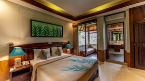 A hotel room with a large bed, wooden headboard, a painting of plants on the wall, a nightstand with a lamp, and a view of the outside through a door. - De Munut Balinese Resort & Spa (Ubud)