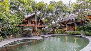 A pool area with lounge chairs and umbrellas, surrounded by lush greenery and traditional architecture. - De Munut Balinese Resort & Spa (Ubud)
