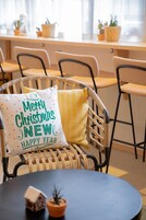 Una silla de mimbre con un cojín que dice 'Merry Christmas New Year' y una pequeña decoración con forma de casa sobre una mesa.
